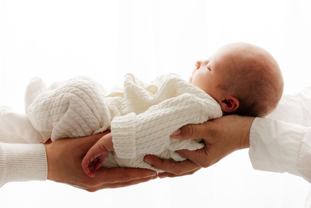 Baby wearing white knit sweater is securely passed between two hands against a white background.