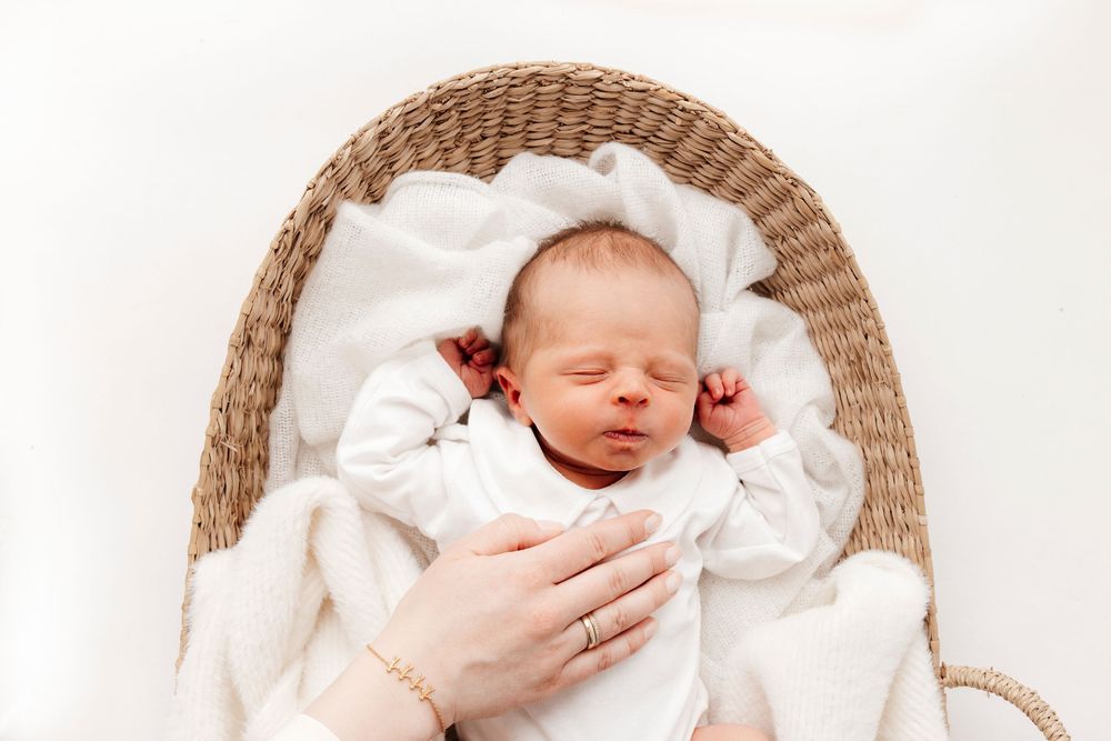 Newborn baby sleeping peacefully in wicker basket lined with soft white blankets while being cradled by gentle hands.
