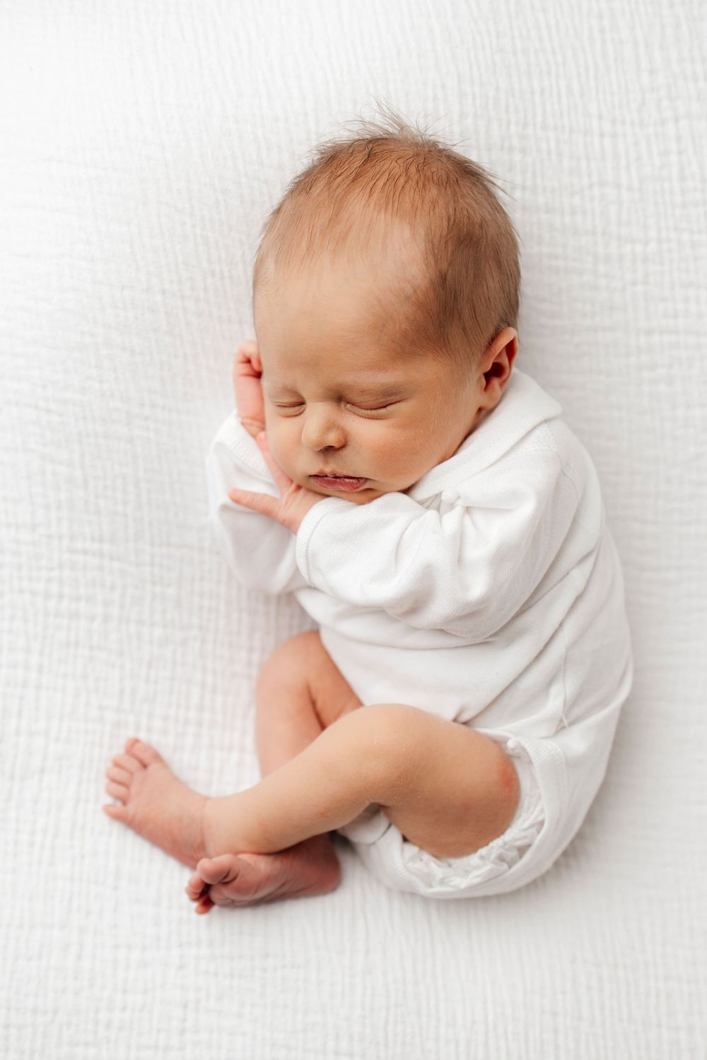 A sleeping newborn baby wearing white clothing lies peacefully on a white textured background.