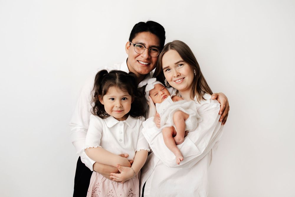 A family of four wearing coordinated white outfits poses together against a light gray studio backdrop.