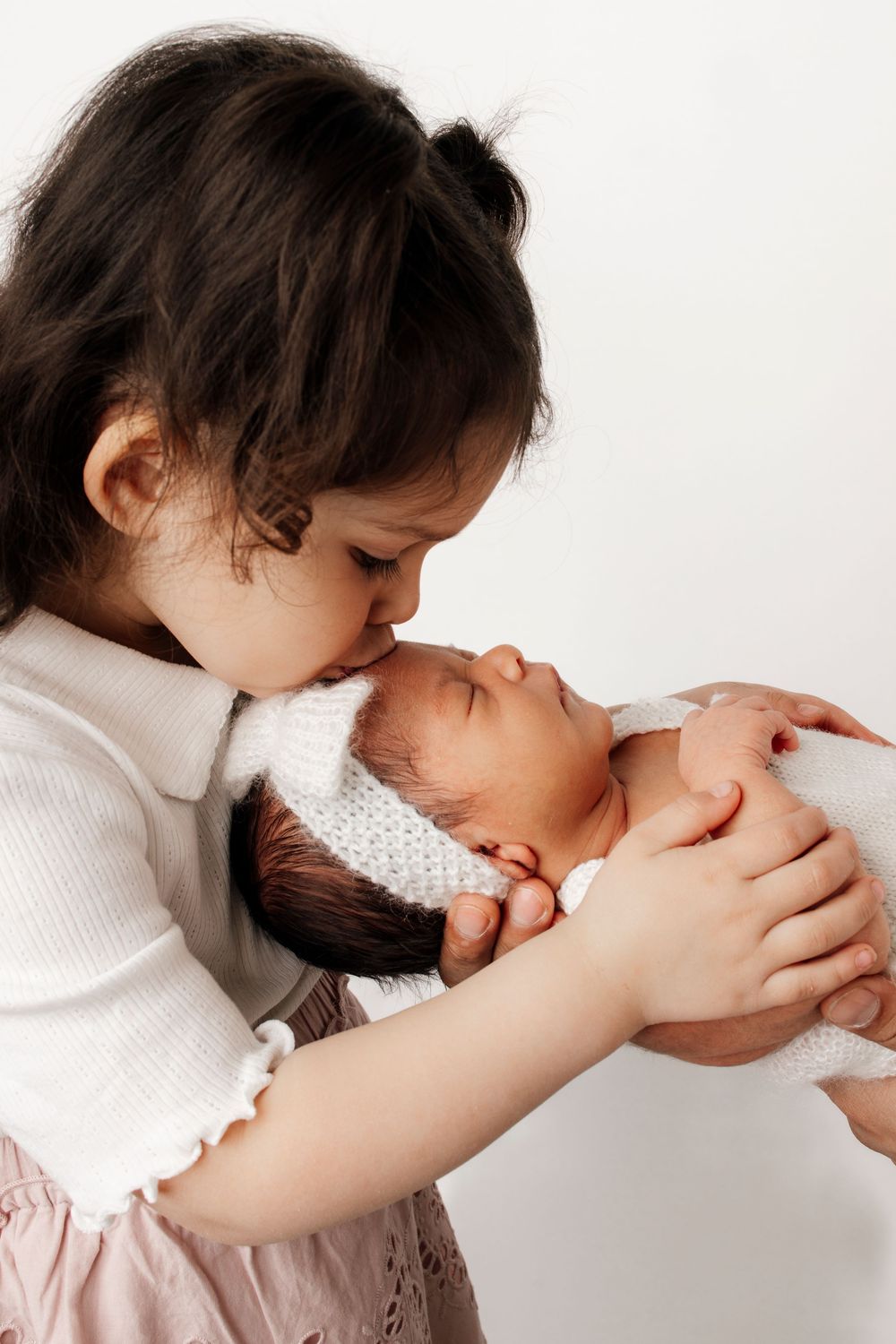 Older sibling wearing white sweater tenderly holds and kisses newborn baby wrapped in knit blanket against white background.