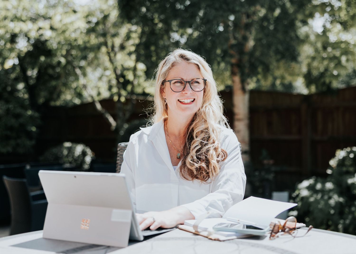 A person in white works on a laptop outdoors while sitting at a wooden table in natural lighting.