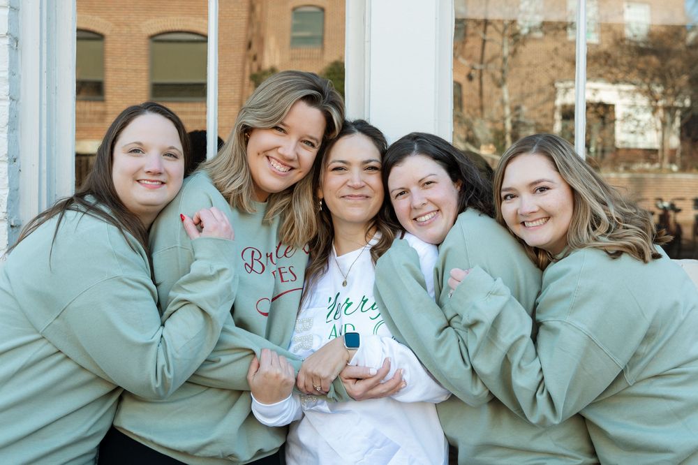 Bride surrounded by bridesmaids wearing matching sweatshirts — candid wedding celebration portrait