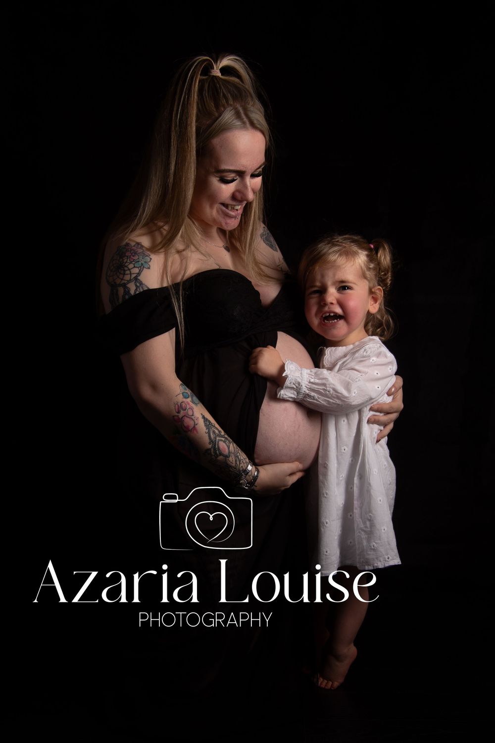 Studio maternity photo shows a pregnant woman in black dress and child in white laughing together against dark background.