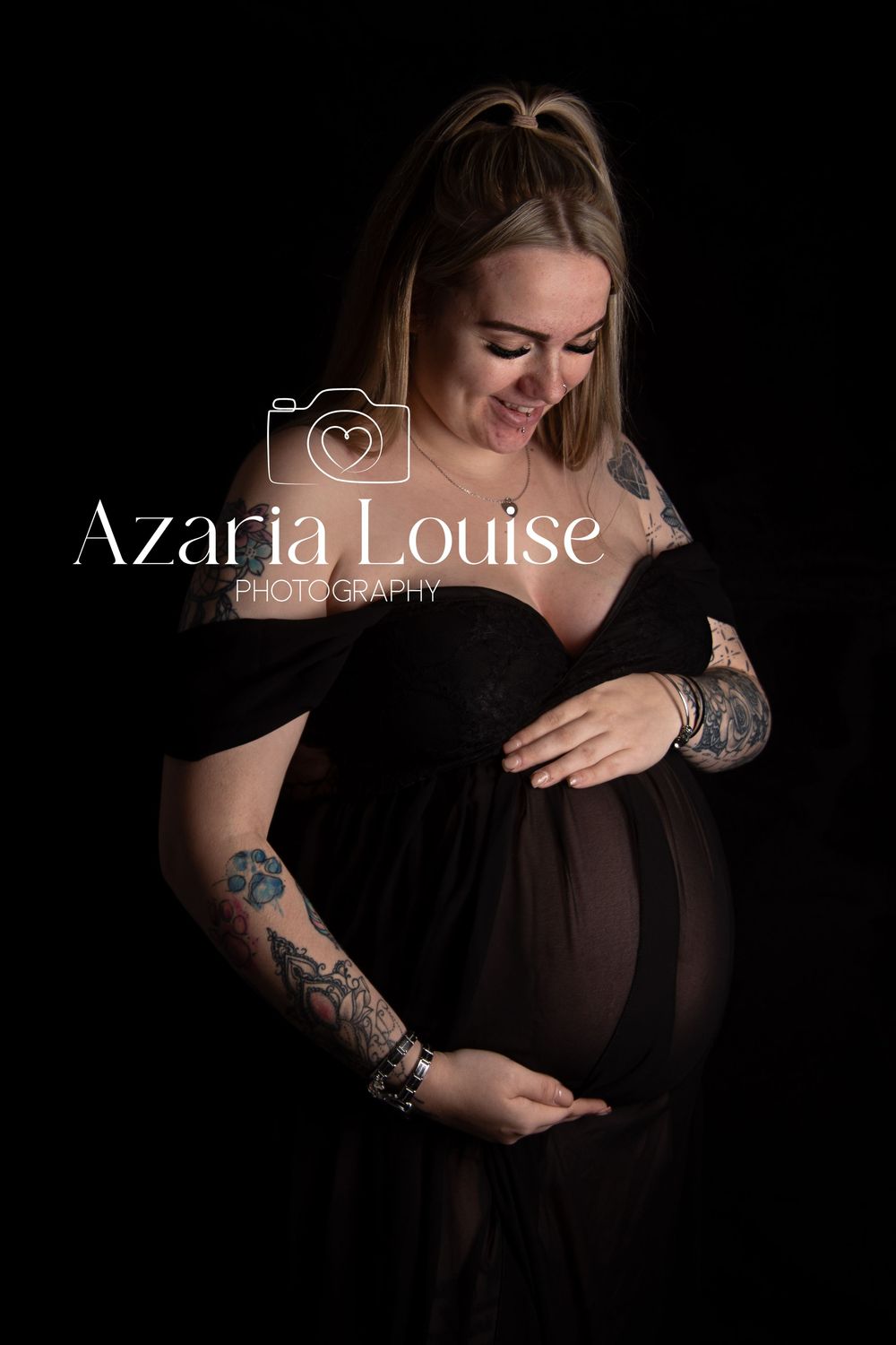 A pregnant person in a black dress poses against dark background with tattoo-covered arms in elegant maternity photo.