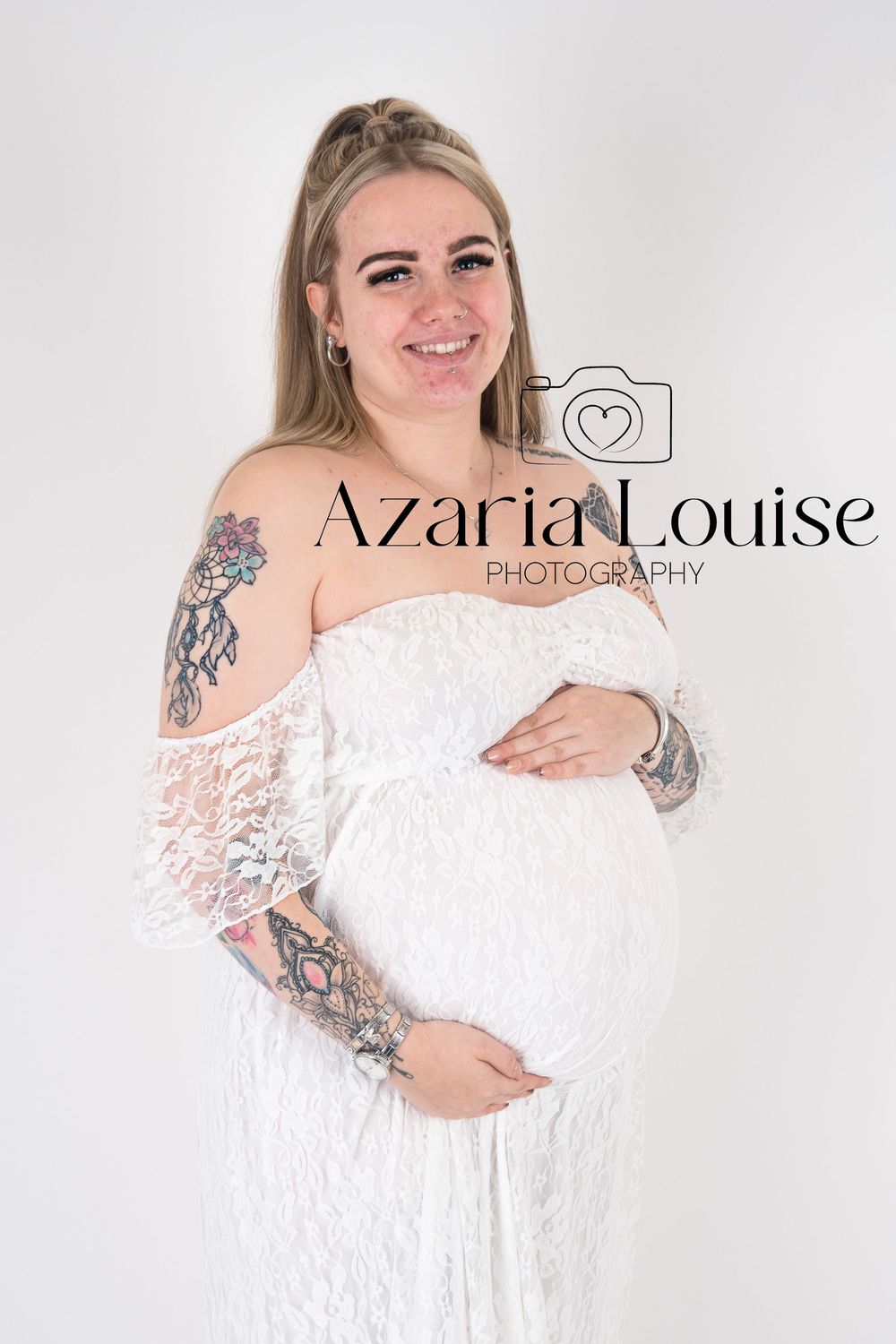 A maternity photo of someone in a white lace off-shoulder dress with visible arm tattoos, smiling against a light background.