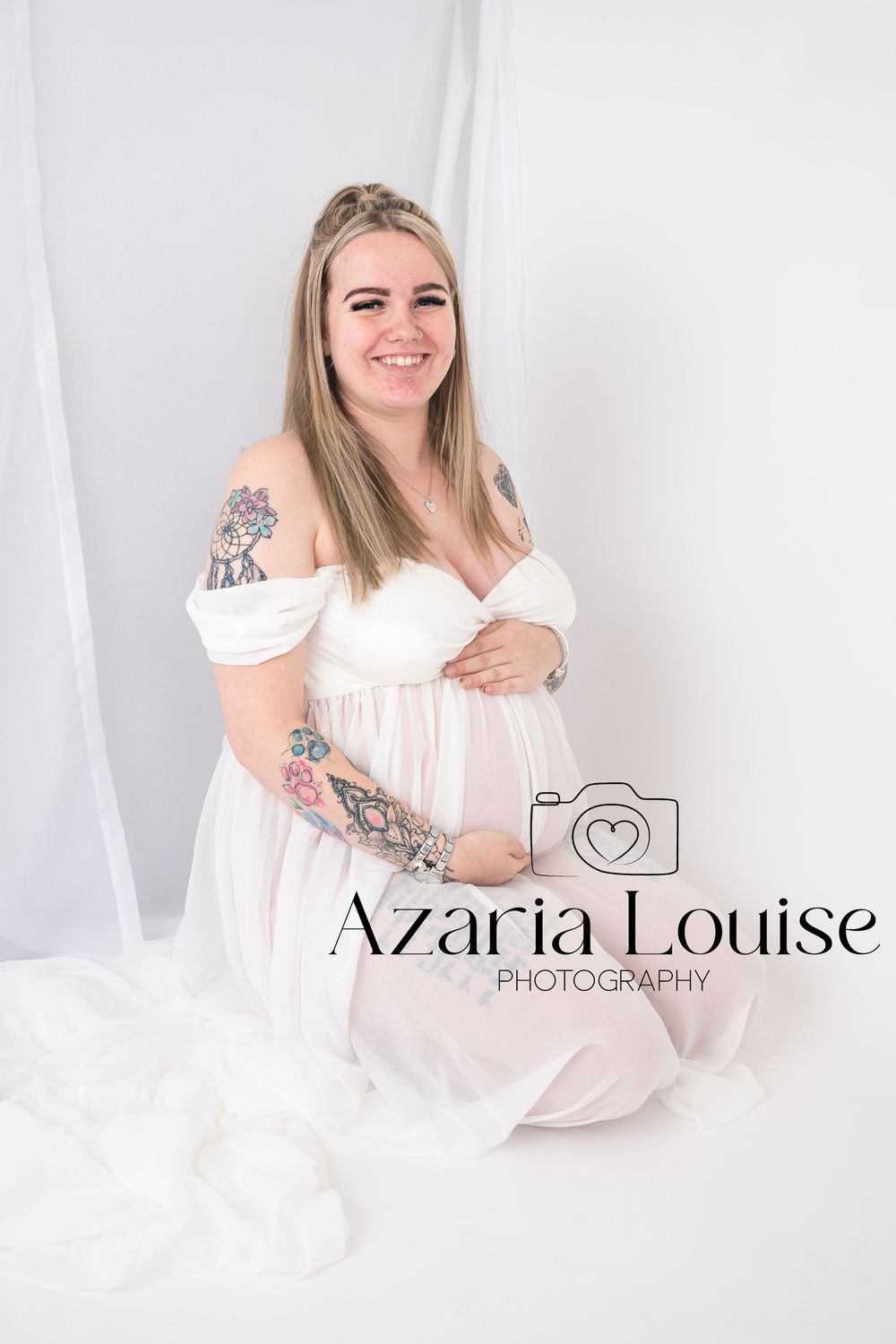 A pregnant woman in a white off-shoulder dress sits on white fabric during a maternity photoshoot with visible tattoos.