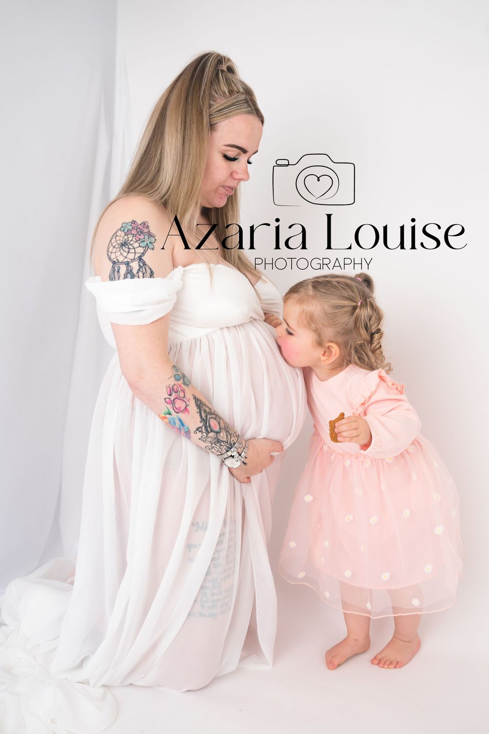 A pregnant woman in a white dress and a small child in pink tulle dress share a tender moment in a minimalist studio setting.
