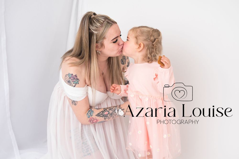 Two people in soft pink and white outfits share a sweet moment against a light background with photographer logo overlay.
