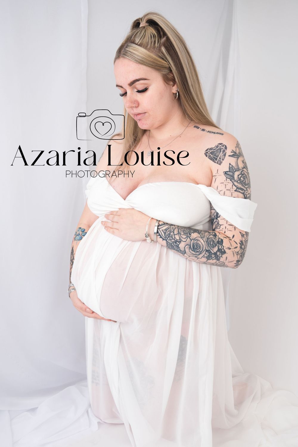 A maternity photo of a person in a white off-shoulder dress with floral tattoo sleeves against a light backdrop.