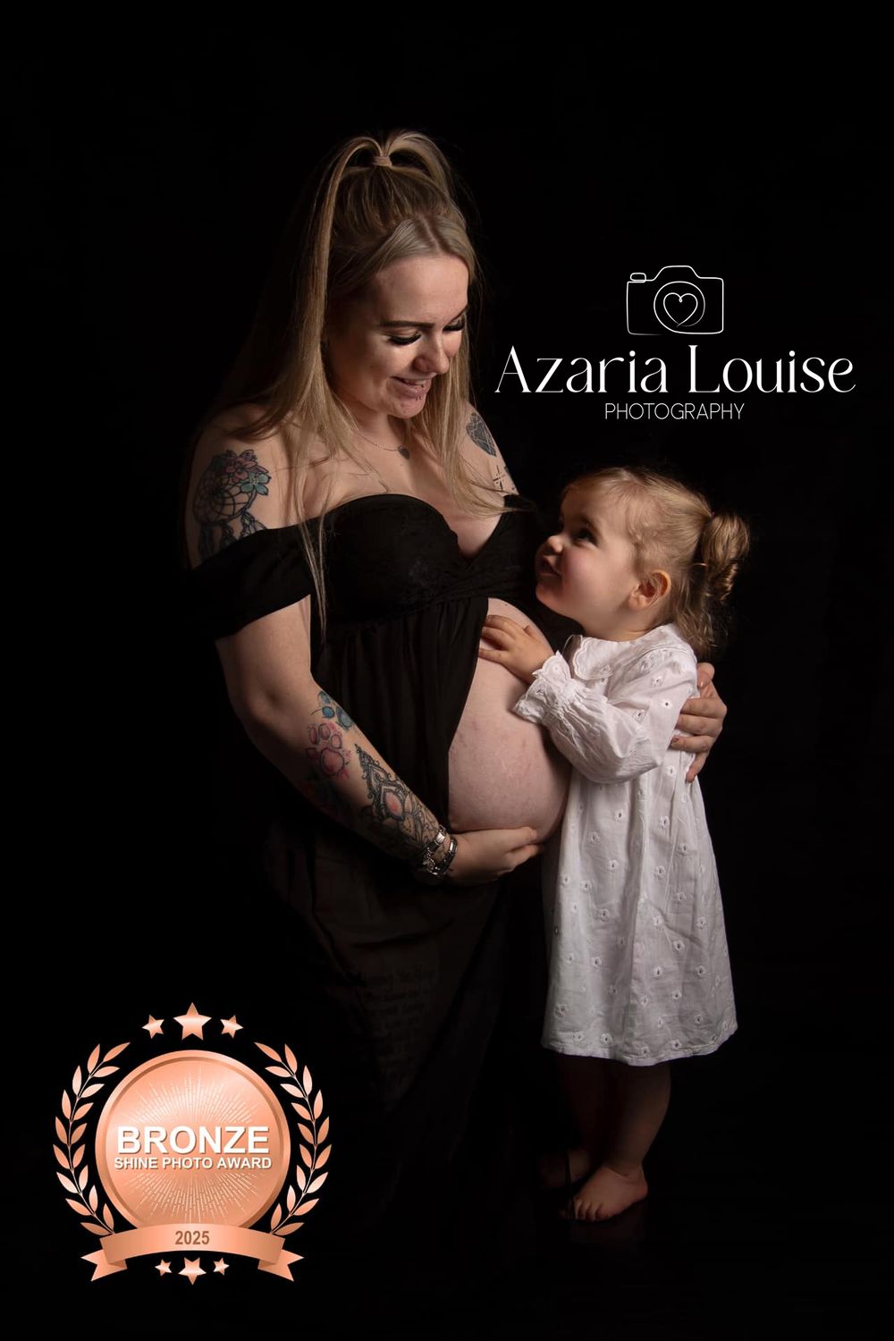 Maternity portrait in black off-shoulder dress with child in white nightgown against dark background, showing Bronze award badge.
