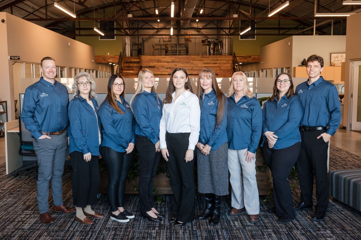 A group of professionals in blue jackets and business attire stand together in an office space for a team photo.
