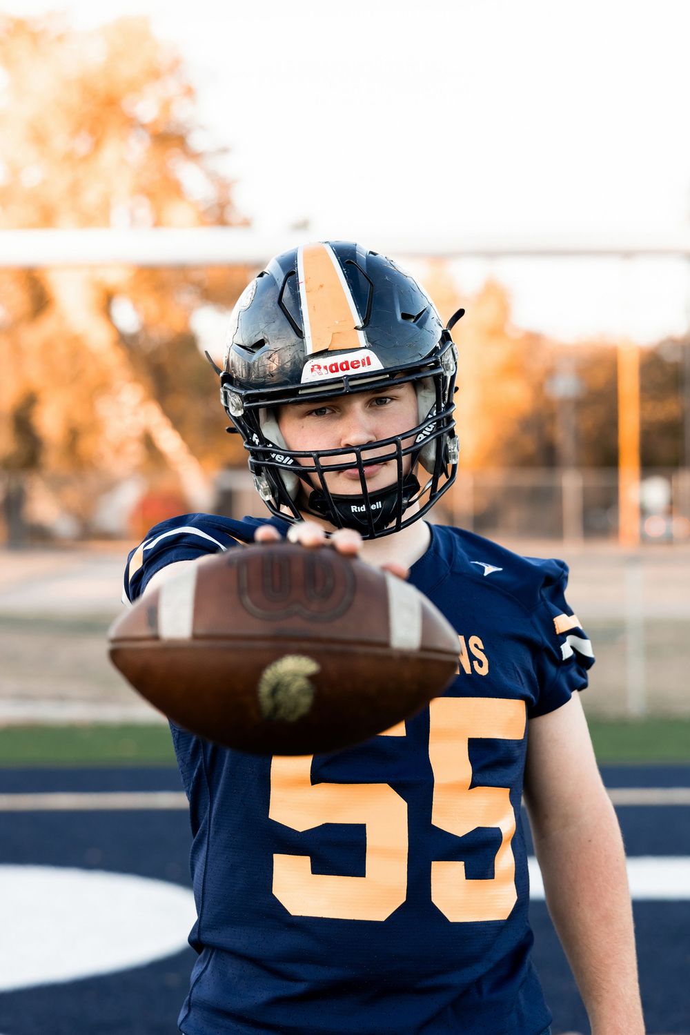 Student athlete in navy blue football uniform number 55 holding a football during golden hour.