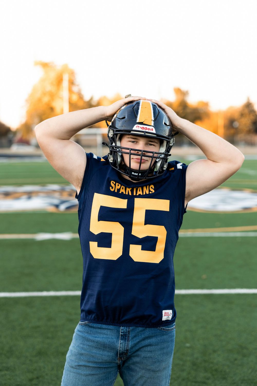 High school football player wearing navy jersey number 55 poses on field at sunset.