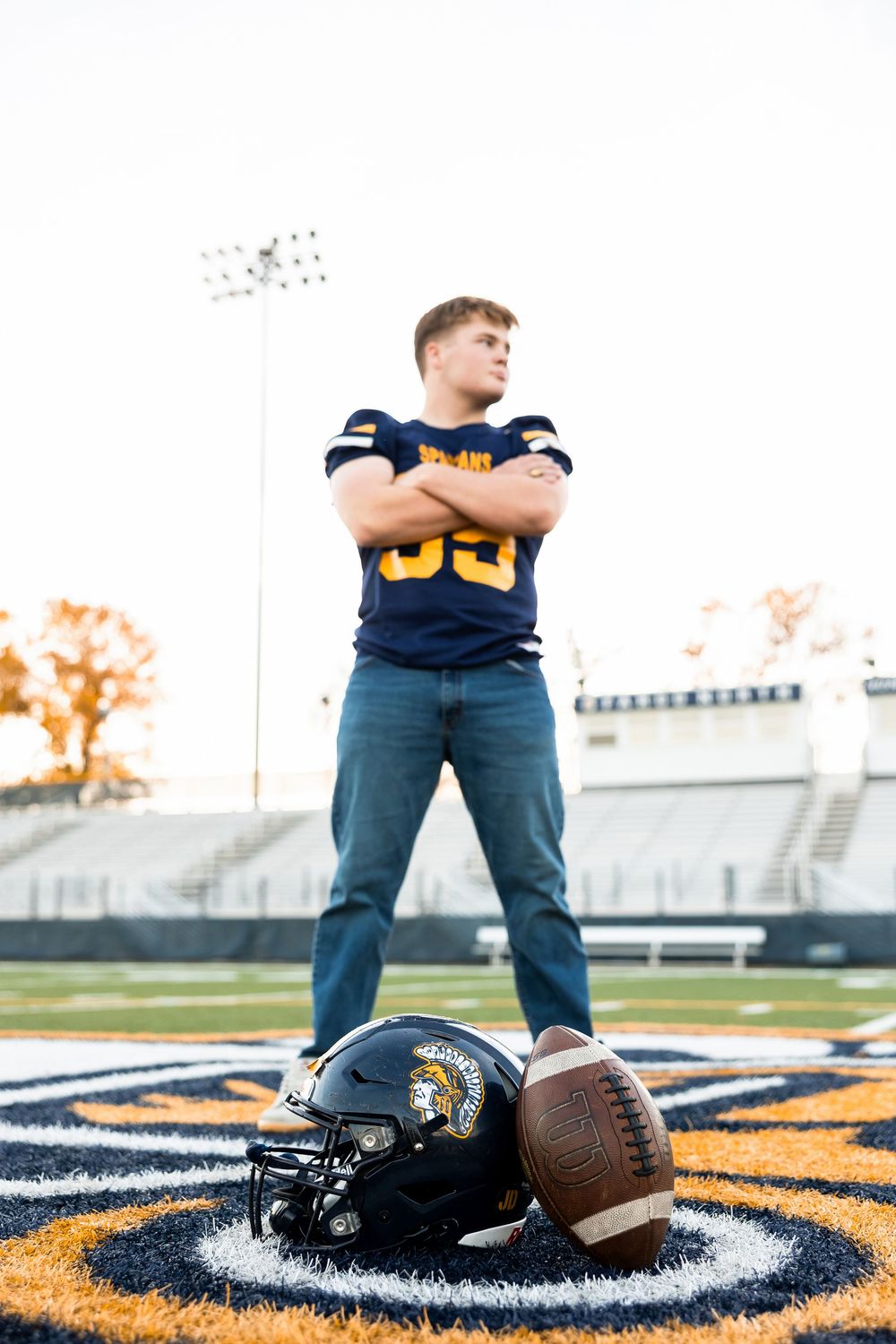 Football player stands on field with helmet and ball at feet while wearing navy jersey and jeans on stadium logo.