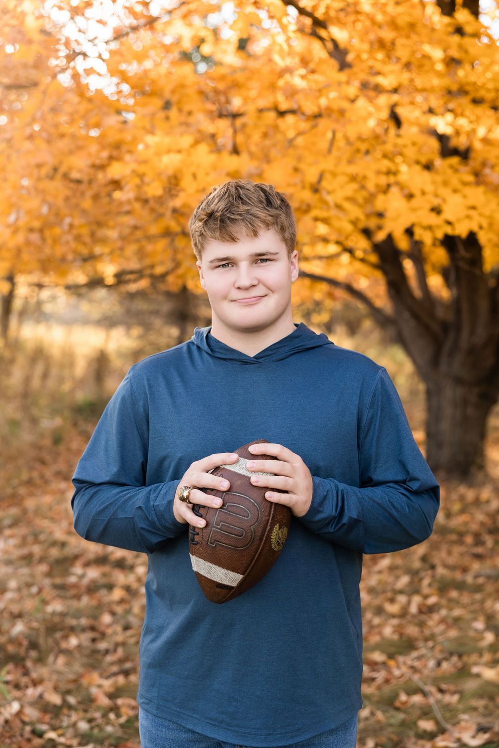 Student athlete poses with football in front of autumn trees with vibrant orange foliage.