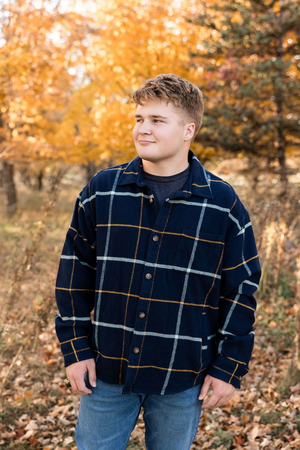 A person wearing a plaid flannel shirt stands in a fall forest with golden autumn foliage in the background.