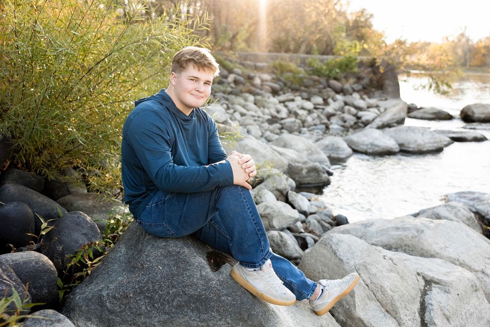 Person relaxing on large rocks along a scenic waterfront during sunset with autumn foliage in background.