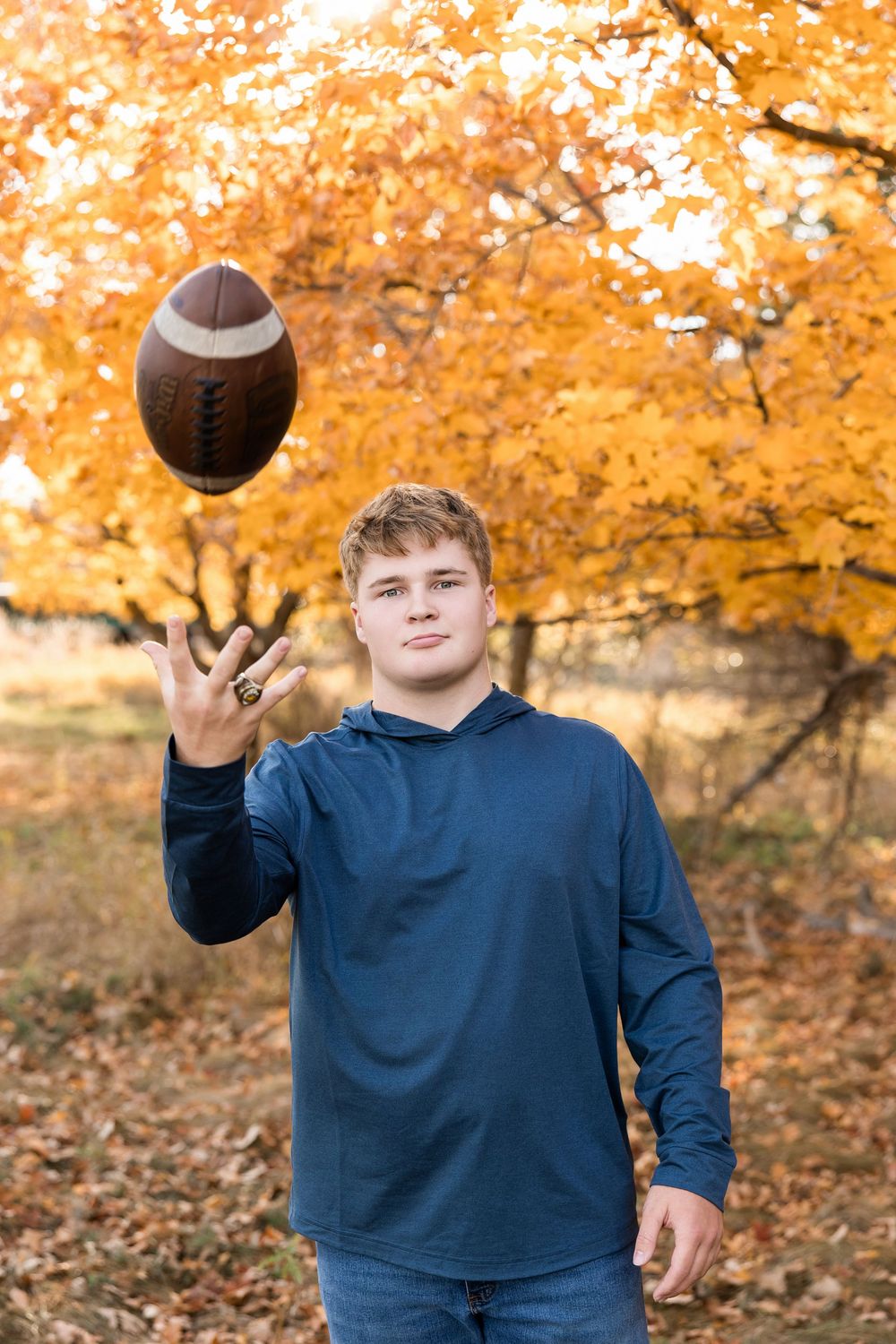A football spins above a person in a navy blue hoodie standing against vibrant fall foliage background.