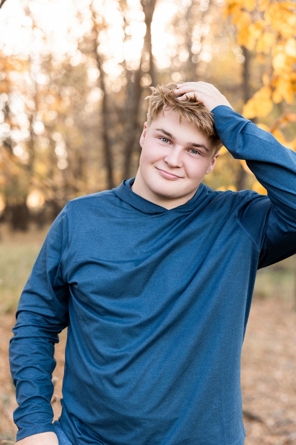 A person in a navy blue sweater poses casually against an autumn forest backdrop with warm lighting.