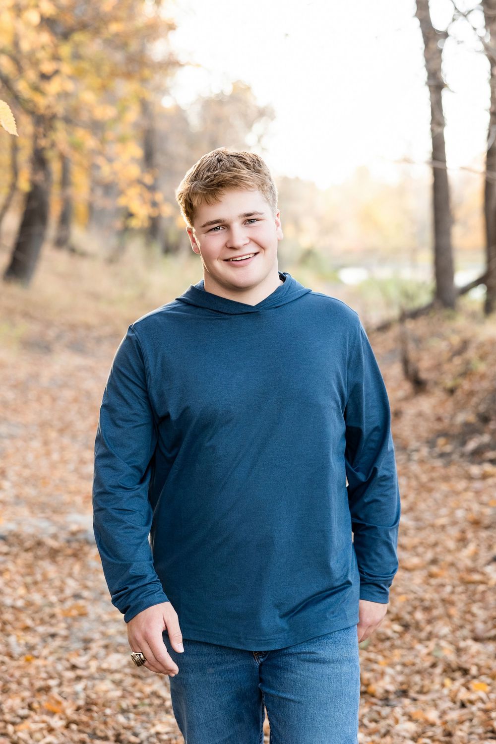A person in a navy hoodie smiles while standing on a leaf-covered path with autumn trees in the background.