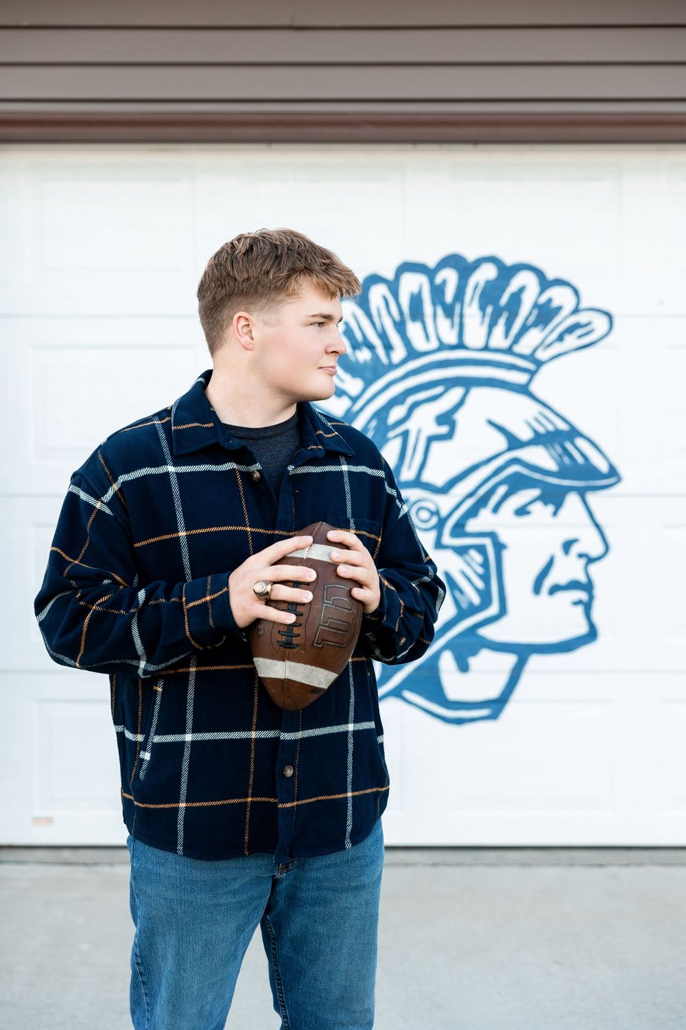 Person in navy plaid shirt holds football while standing against wall with blue trojan mascot graphic in background.