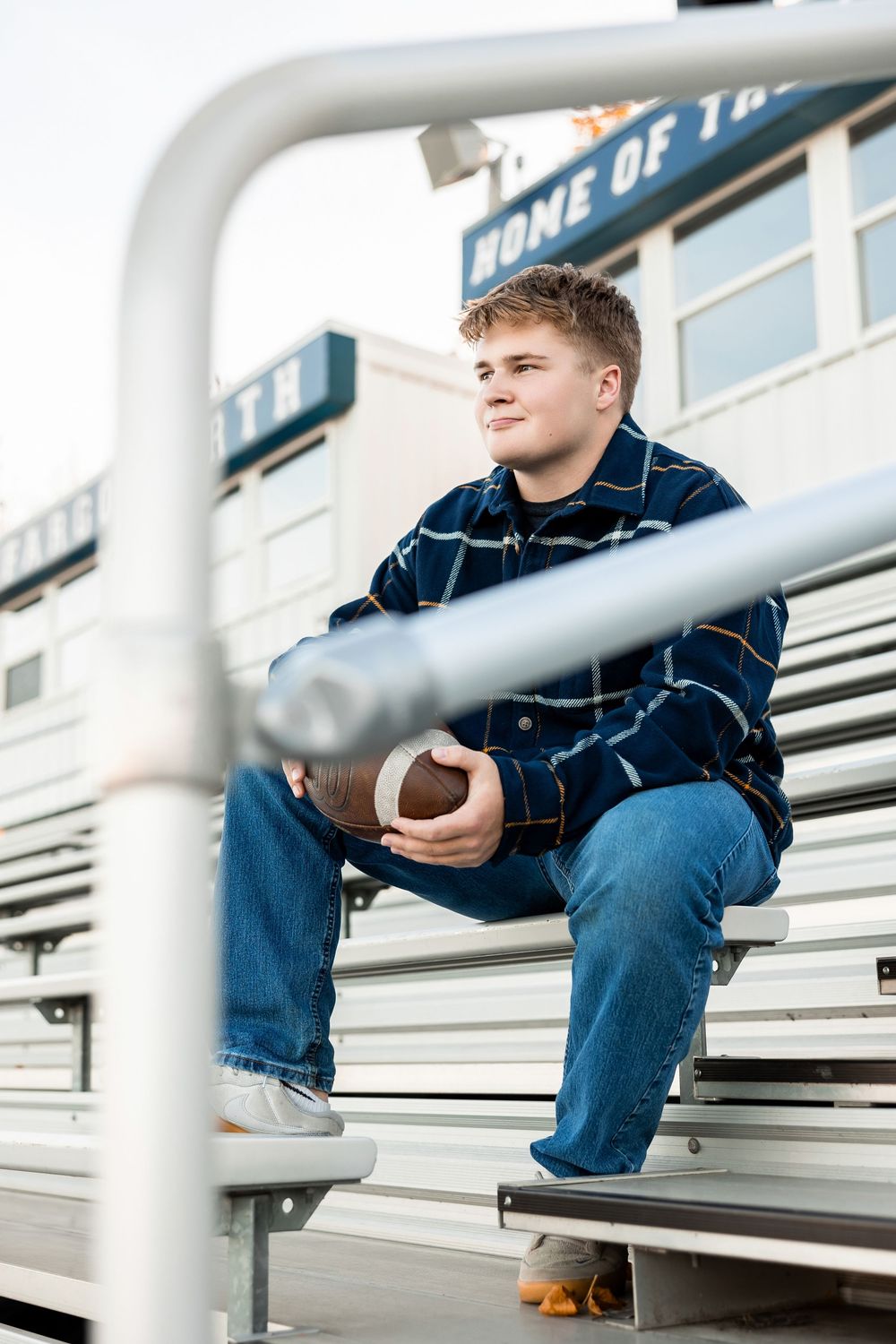 Football player sits on metal bleachers holding football at stadium.