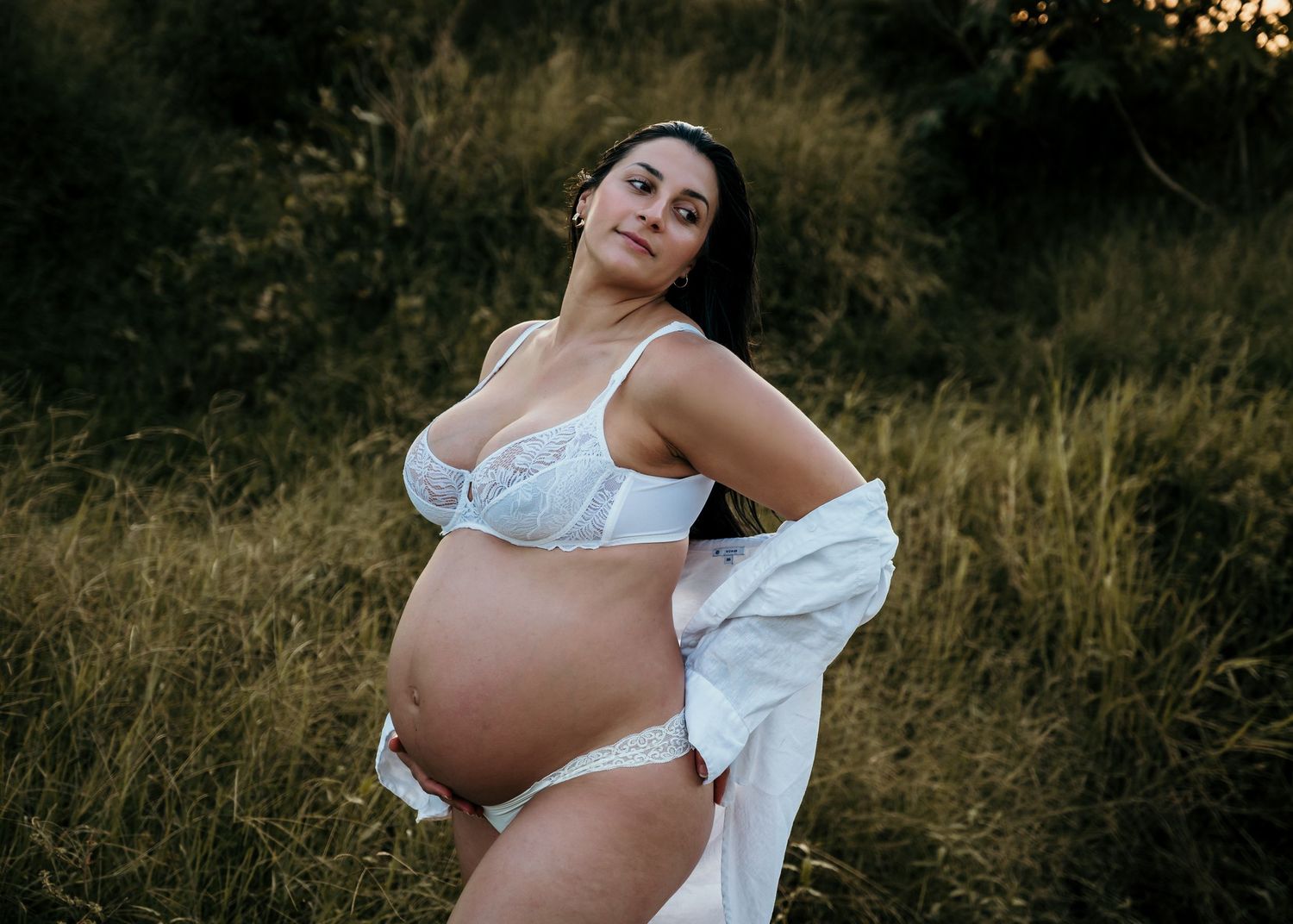 Expectant mother in white lingerie poses confidently in golden hour sunlit field.
