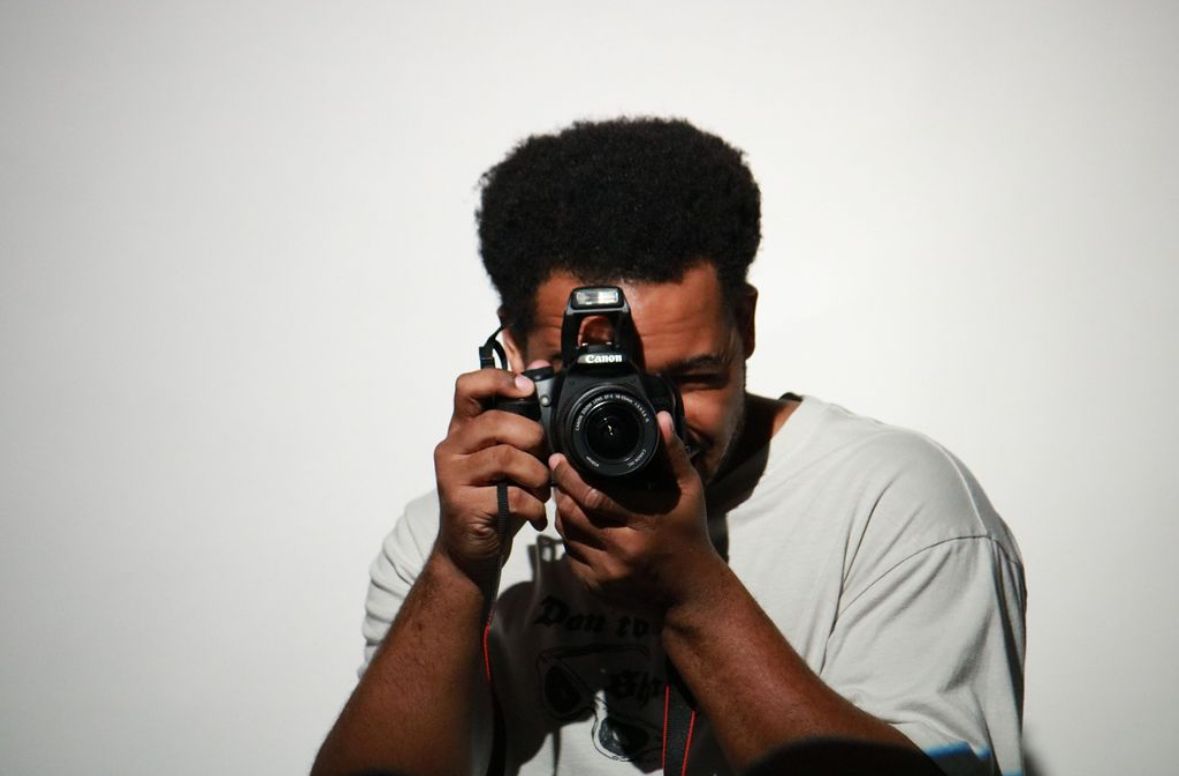 Photographer in casual attire taking pictures with professional camera against light background.