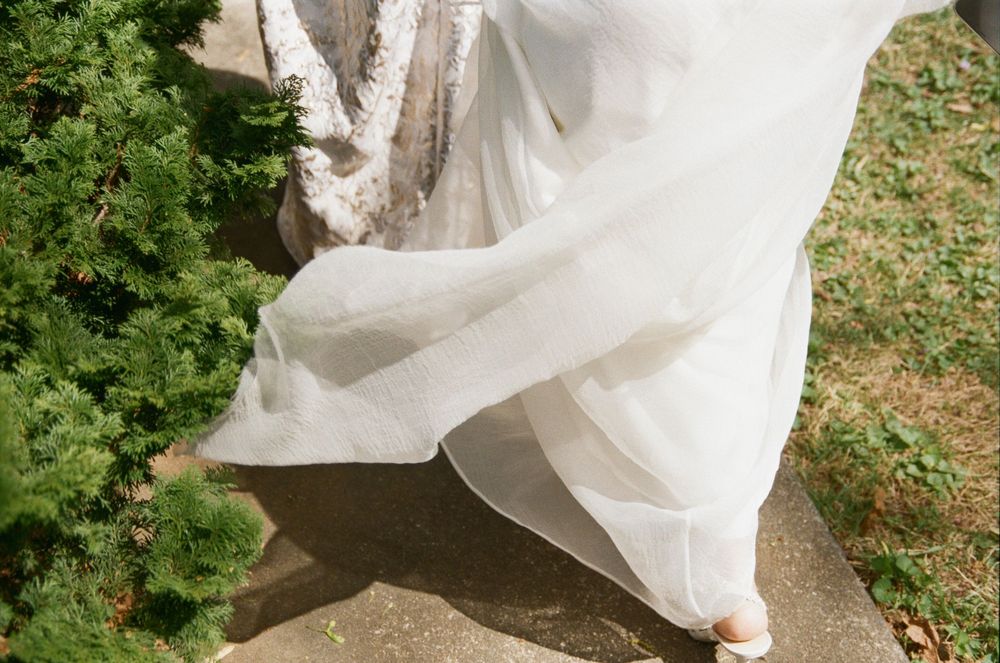 A flowing white wedding dress fabric billows gracefully in the breeze against green foliage background.