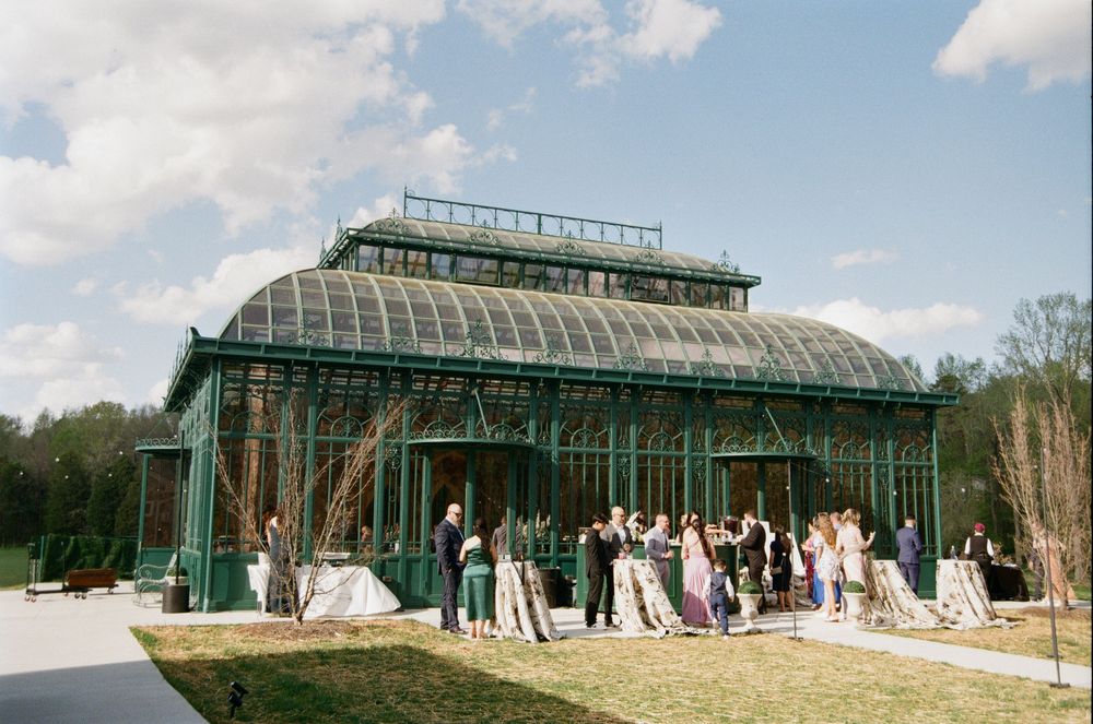 A vintage-style glass and steel greenhouse venue with guests gathered outside during a special event.