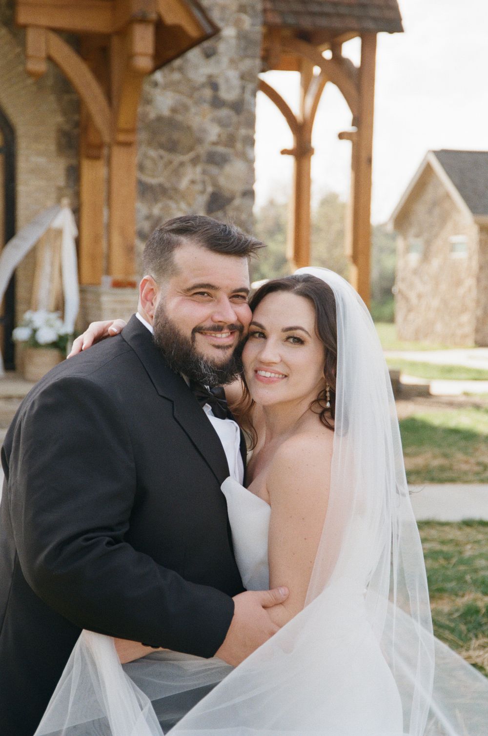 A happy couple in formal wedding attire embracing outdoors under a wooden pergola with stone architecture.