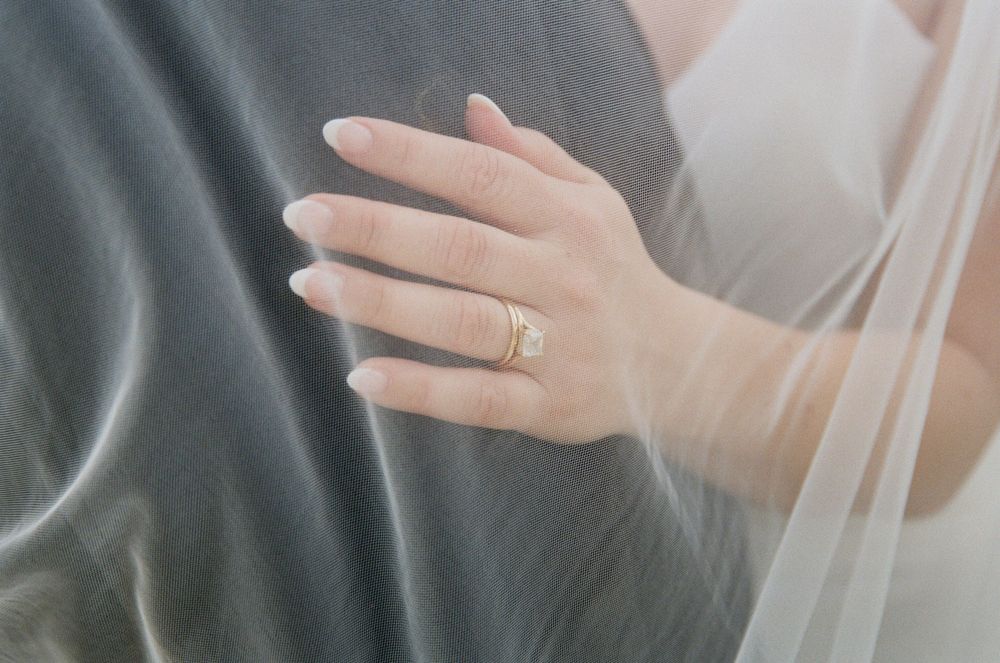 A delicate hand wearing an engagement ring rests on a gray fabric, with soft white veil material flowing in the frame.