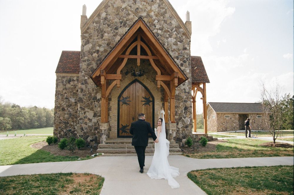 Two people enter a rustic stone chapel with wooden beam details and arched doorway in a countryside setting.