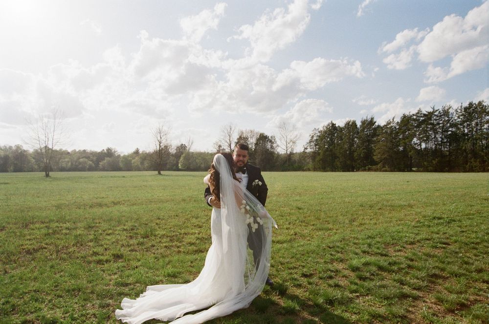 A bride in a flowing white gown and veil stands with their partner in a lush green field on a sunny day.