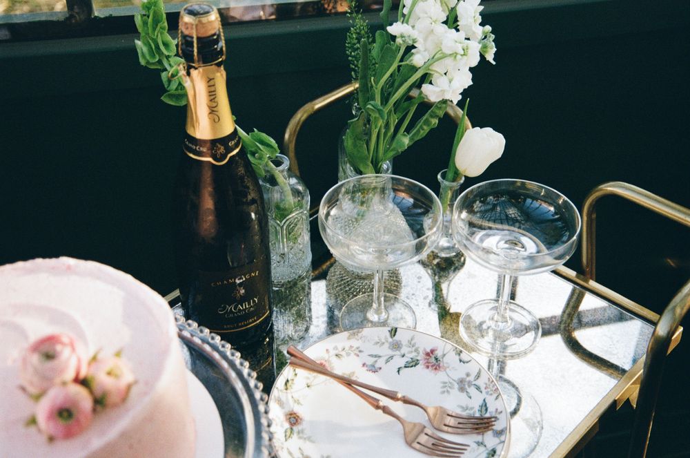 Elegant party setup with champagne bottle, crystal glasses, and white flowers in vases on a mirrored tray.