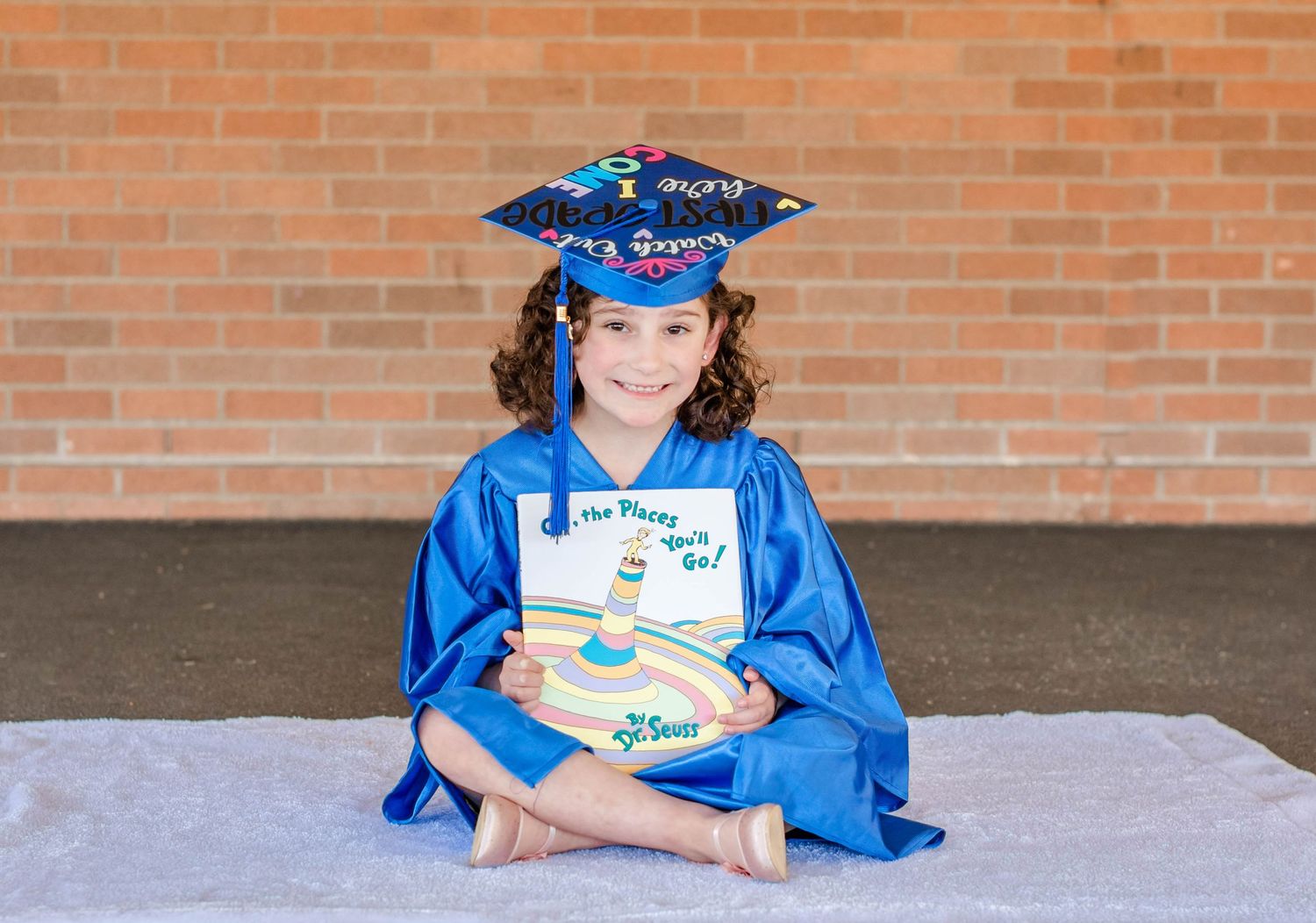 A graduate in a bright blue cap and gown poses against a brick wall background.