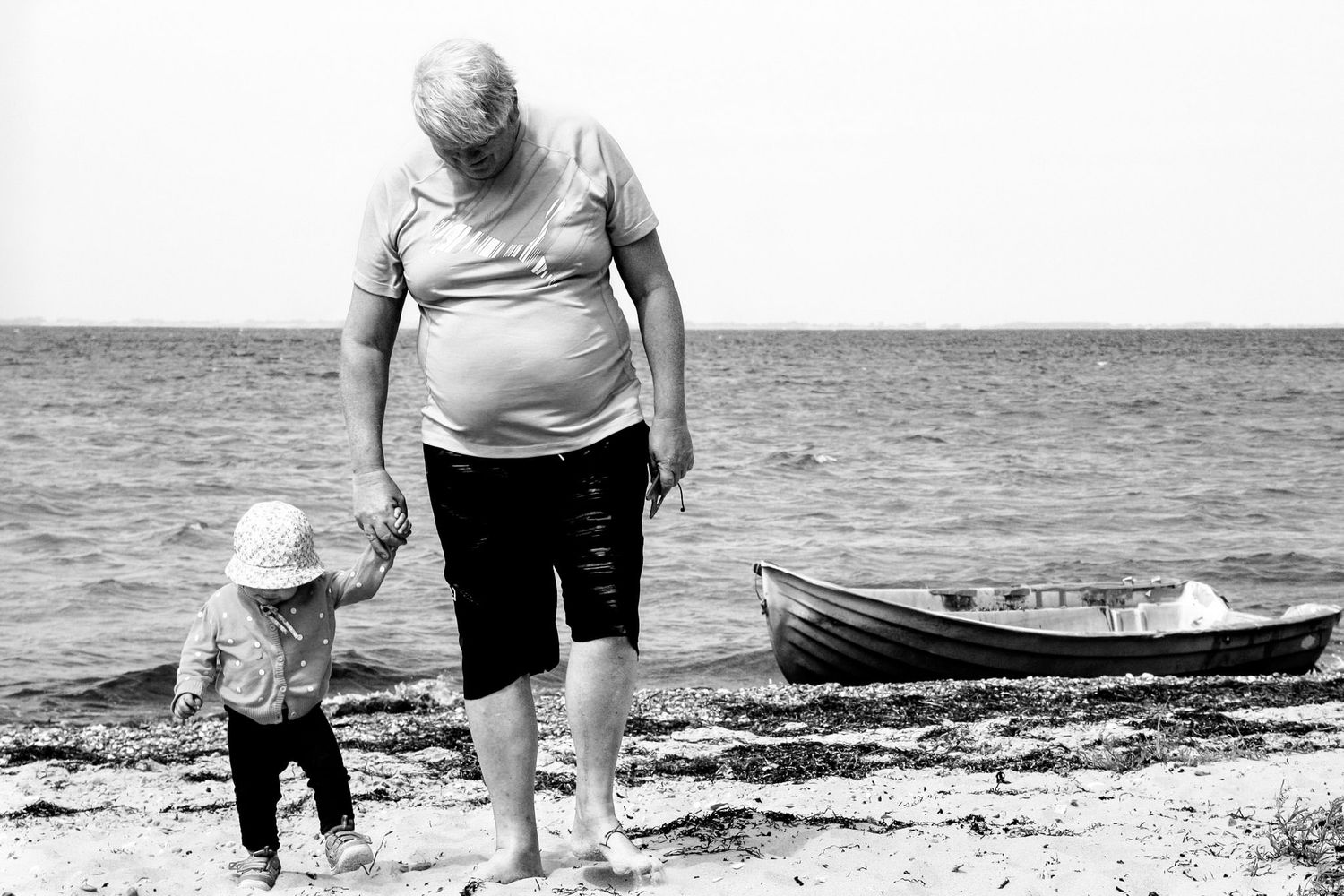 En morfar og et barnebarn som går hånd i hånd på en strand med en gammel båt i bakgrunnen. Tatt av familiefotograf Tone Ørstavik i Rogaland.