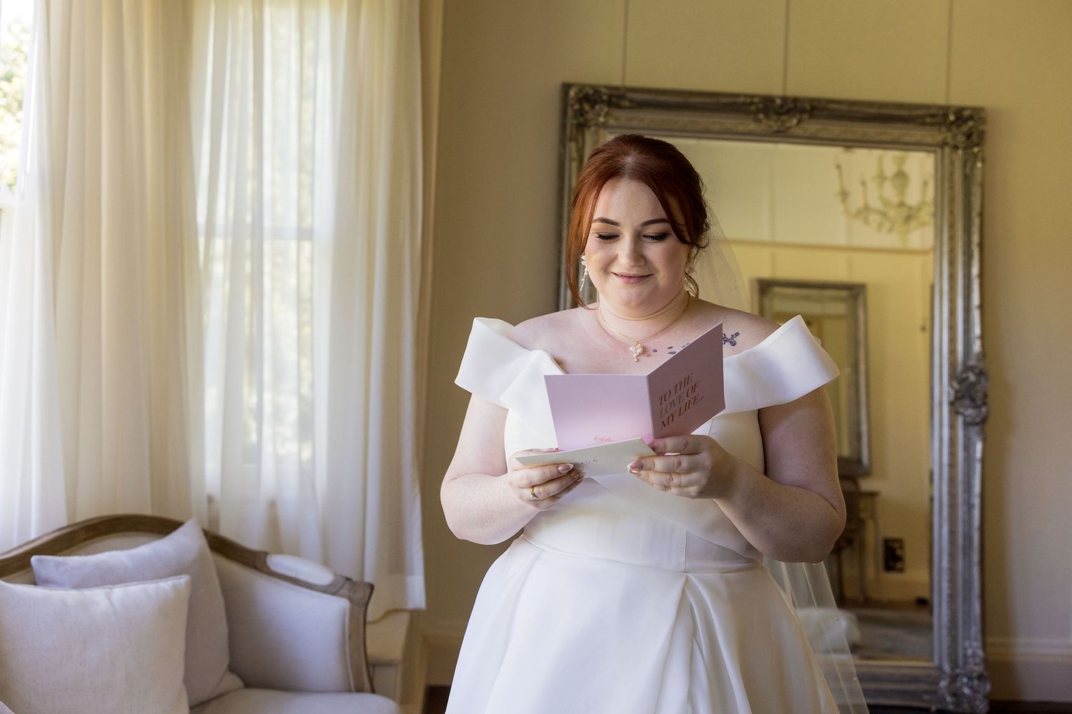 Bride in a white wedding dress holds a letter from the Groom while standing in an elegant room with sheer curtains at Gabbinbar Homestead