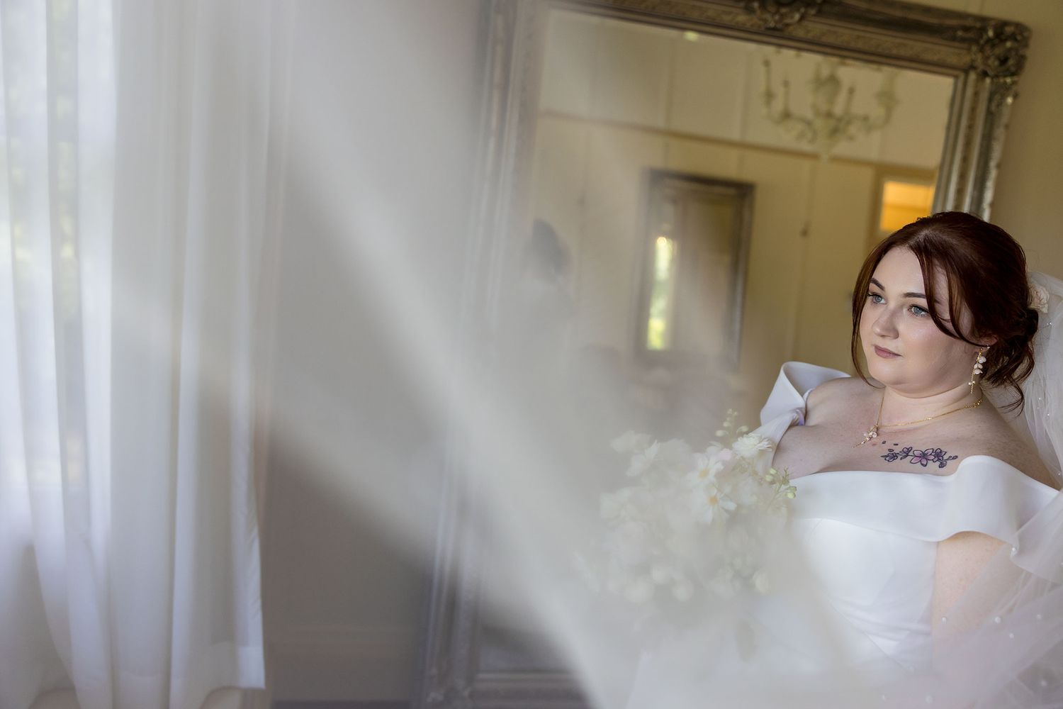 Bride in a white wedding dress holds a bouquet of white flowers while standing in an elegant room with sheer curtains at Gabbinbar Homestead
