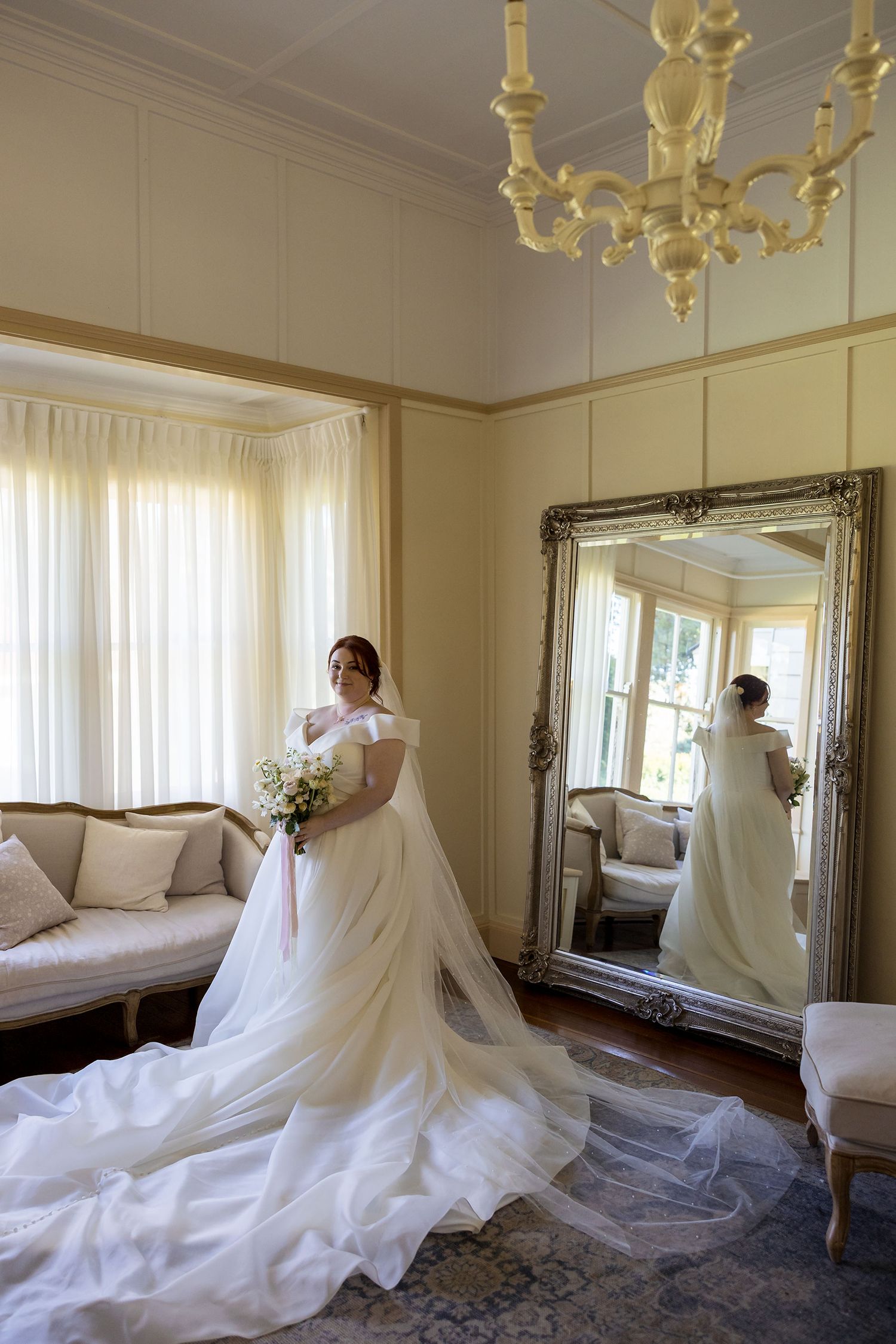 Bride in a white wedding dress holds a bouquet of white flowers while standing in an elegant room with sheer curtains at Gabbinbar Homestead