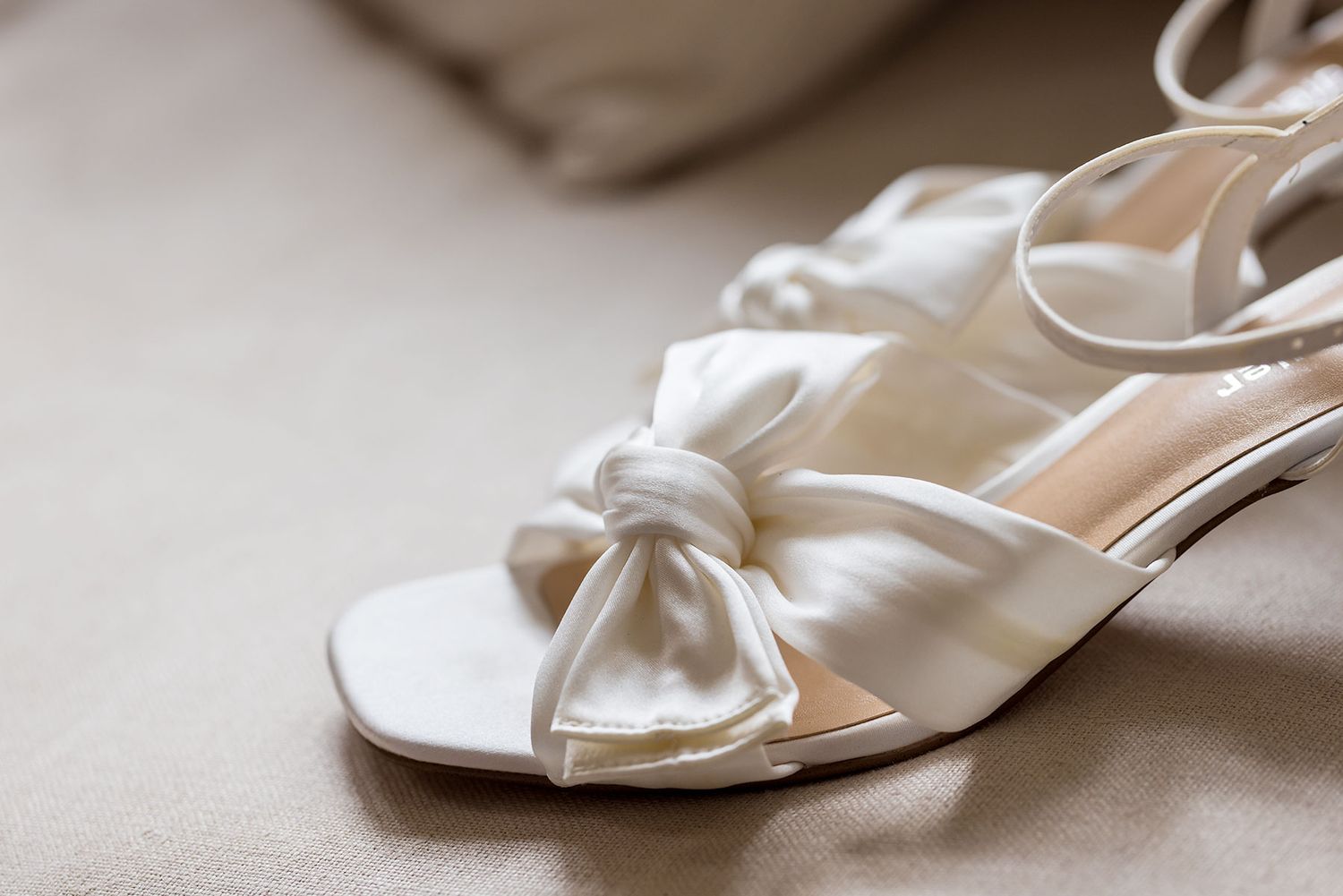 White satin bridal sandals with bow detail sitting on beige fabric background at Gabbinbar Homestead in Toowoombar Queensland.