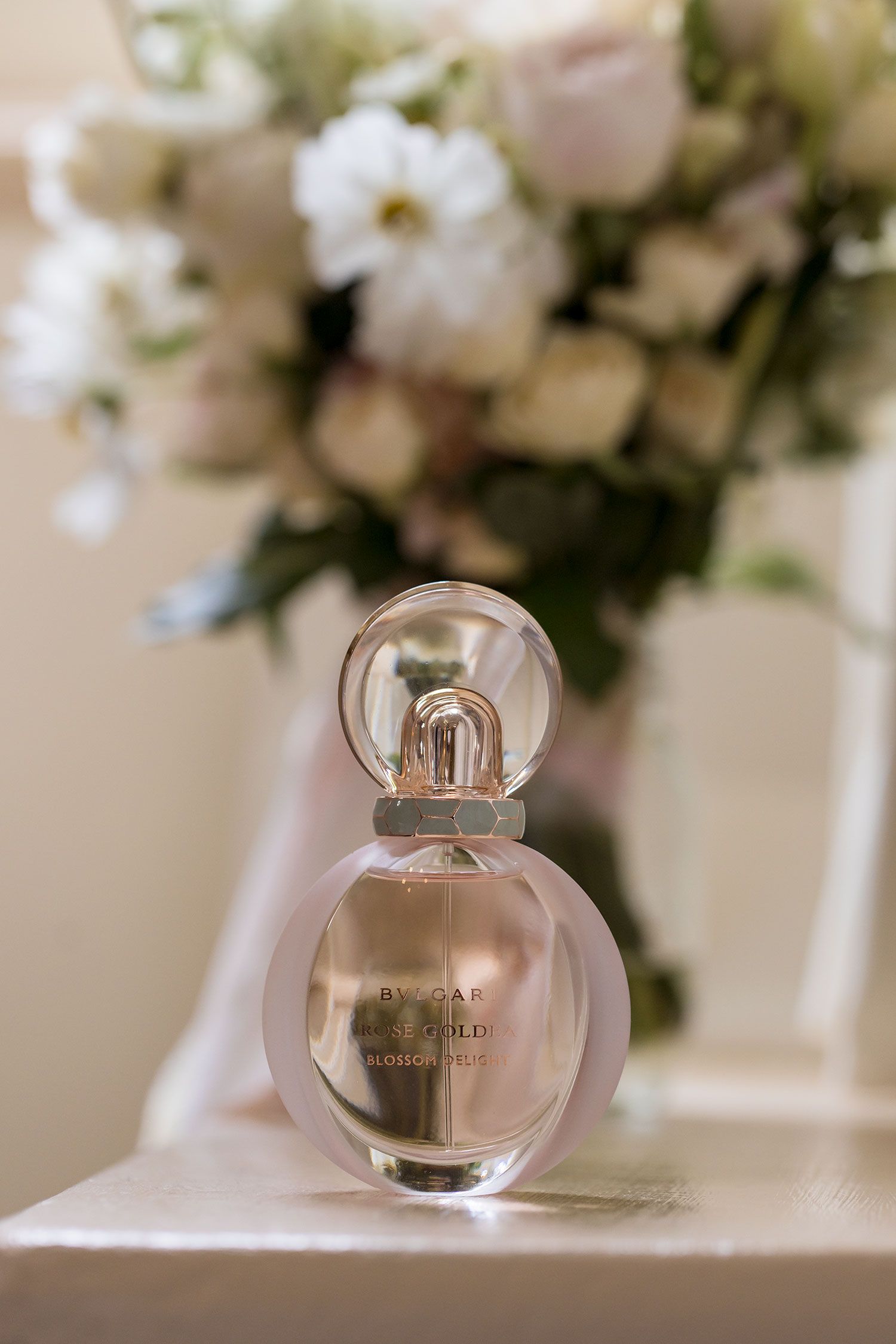 A round glass perfume bottle sits elegantly against a blurred background of soft white flowers and pink roses during wedding preparations.