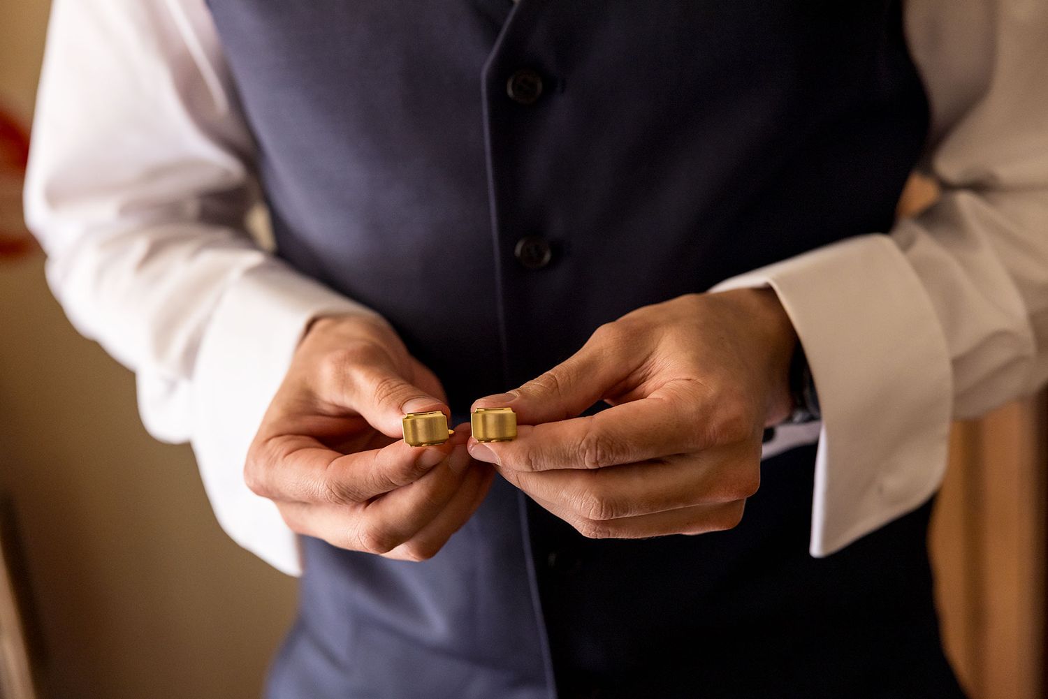 A close up image of a Groom in his blue wedding suit holding his gold cuff links against a warm-lit background at Gabbinbar Toowoomba.