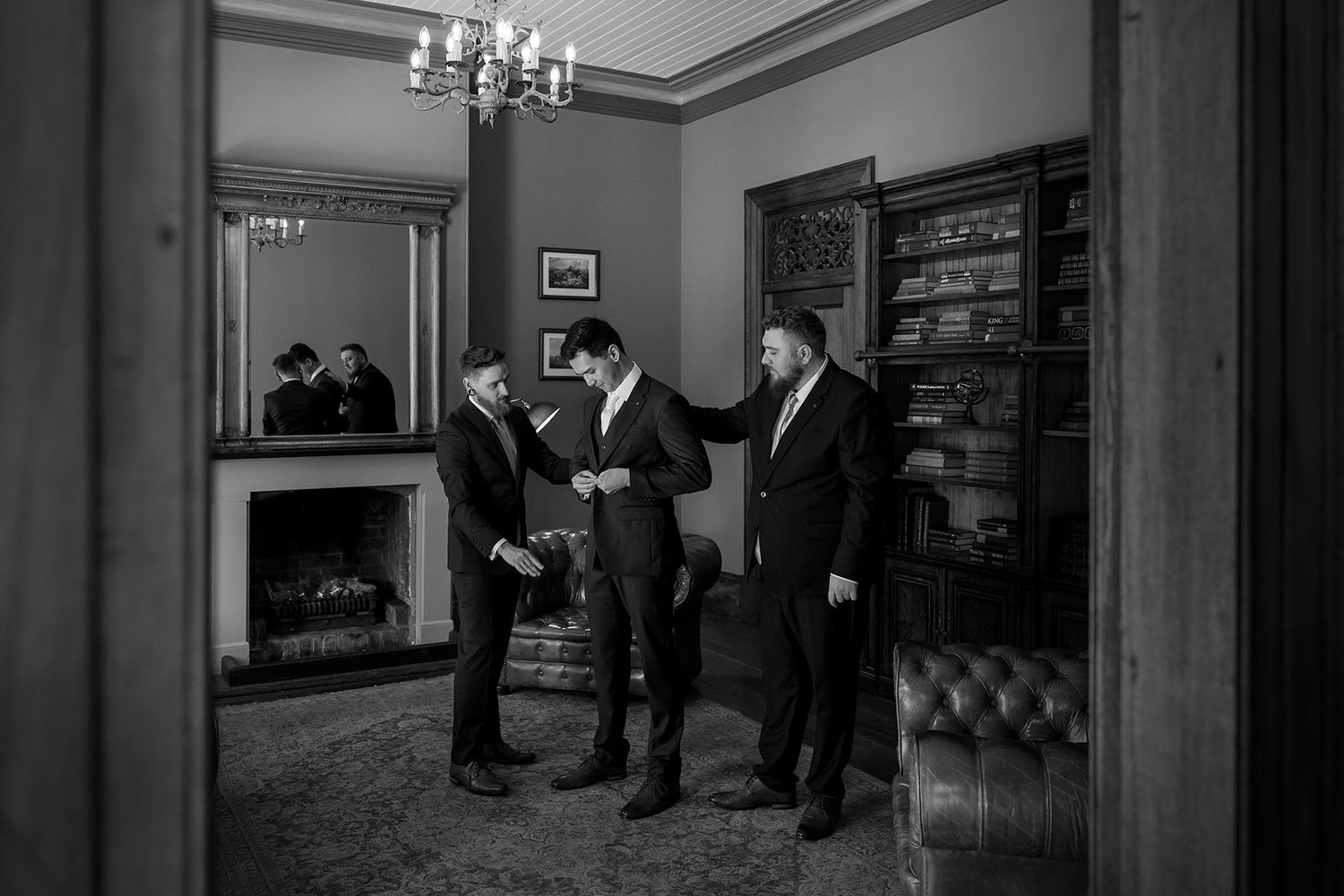 Three men in formal suits stand in an elegant room with a chandelier and fireplace having a discussion.