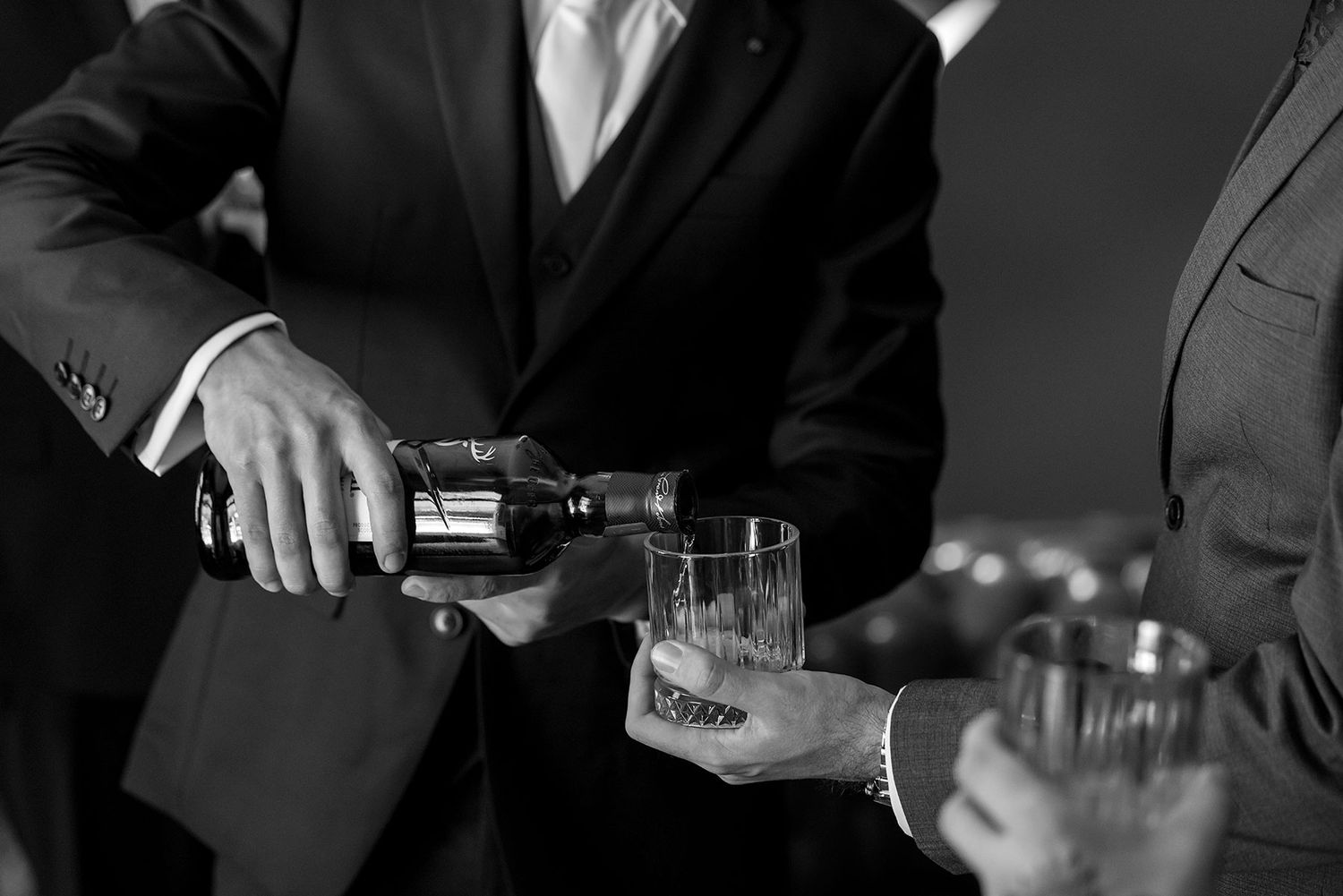 A Groom in a suit pours whiskey into a Groomsman's glass during wedding preparations in a black and white moment at Gabbinbar Homestead.