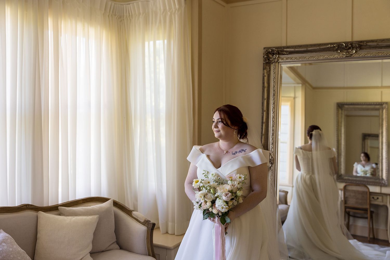 Bride in a white wedding dress holds a bouquet of white flowers while standing in an elegant room with sheer curtains at Gabbinbar Homestead