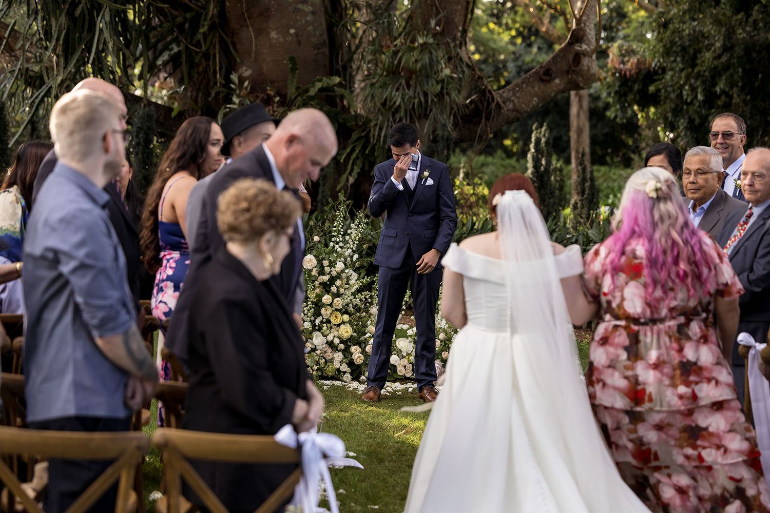 Outdoor garden wedding ceremony with guests gathered around bride in flowing white dress and veil walking towards her Husband to be.