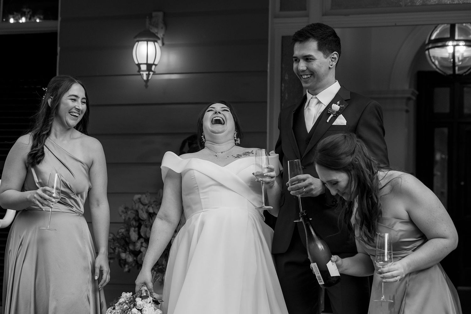 A joyful wedding party shares a laugh together on a porch illuminated by outdoor lanterns on the veranda at Gabbinbar Homestead in Toowoomba