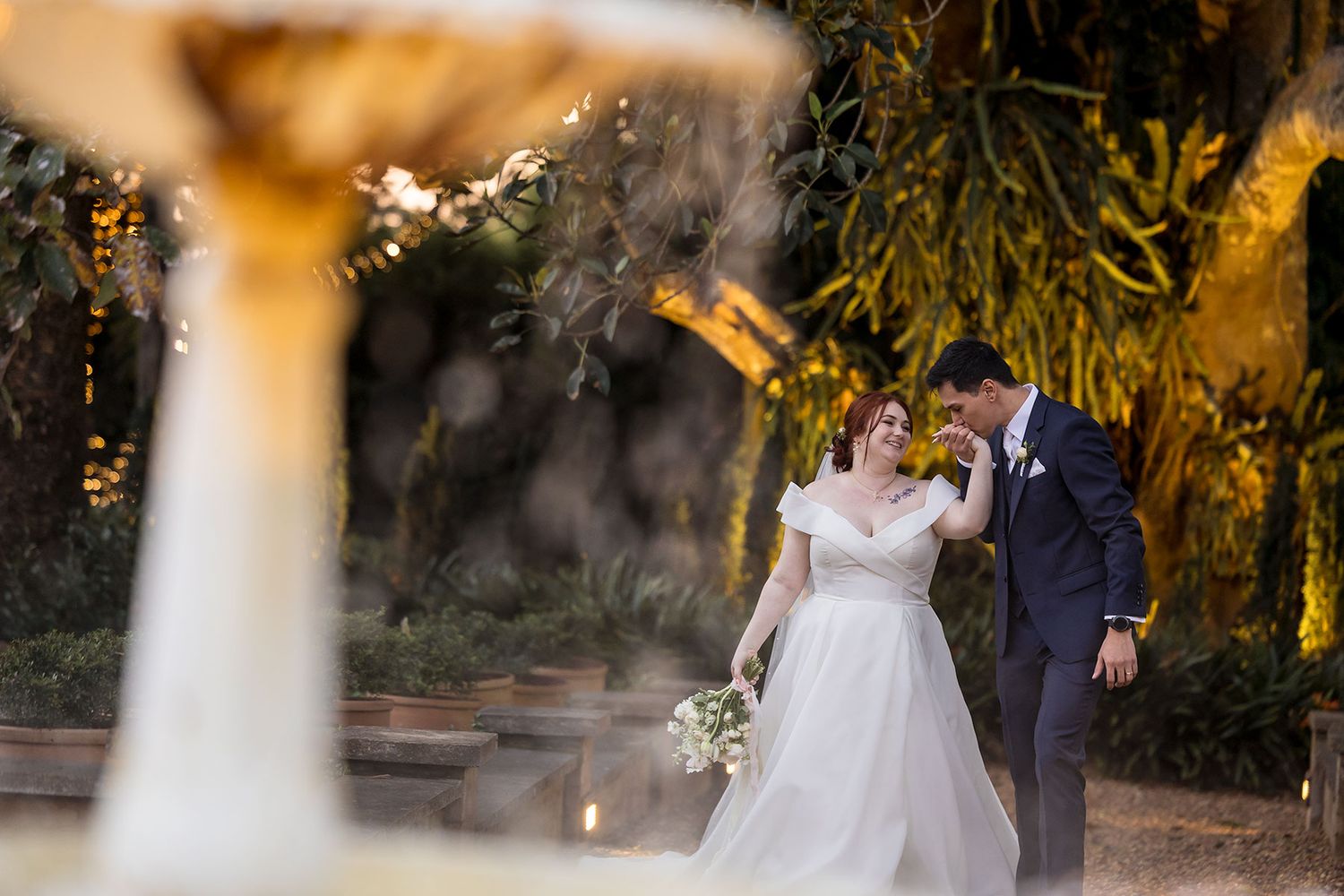 Groom kisses his Bride on the hand in a intimate moment near a water fountain in the gardens at Gabbinbar Homestead Toowoombar Queensland.